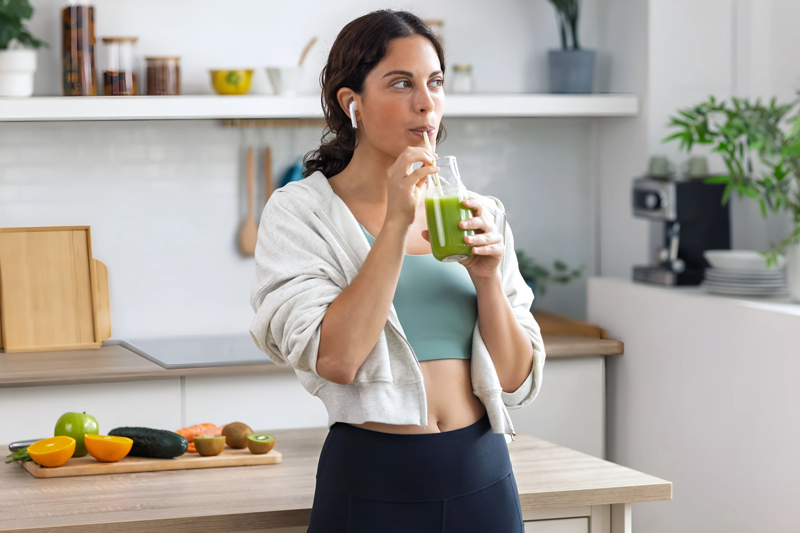 Woman in a kitchen drinking a green smoothie, with fresh fruit on the counter