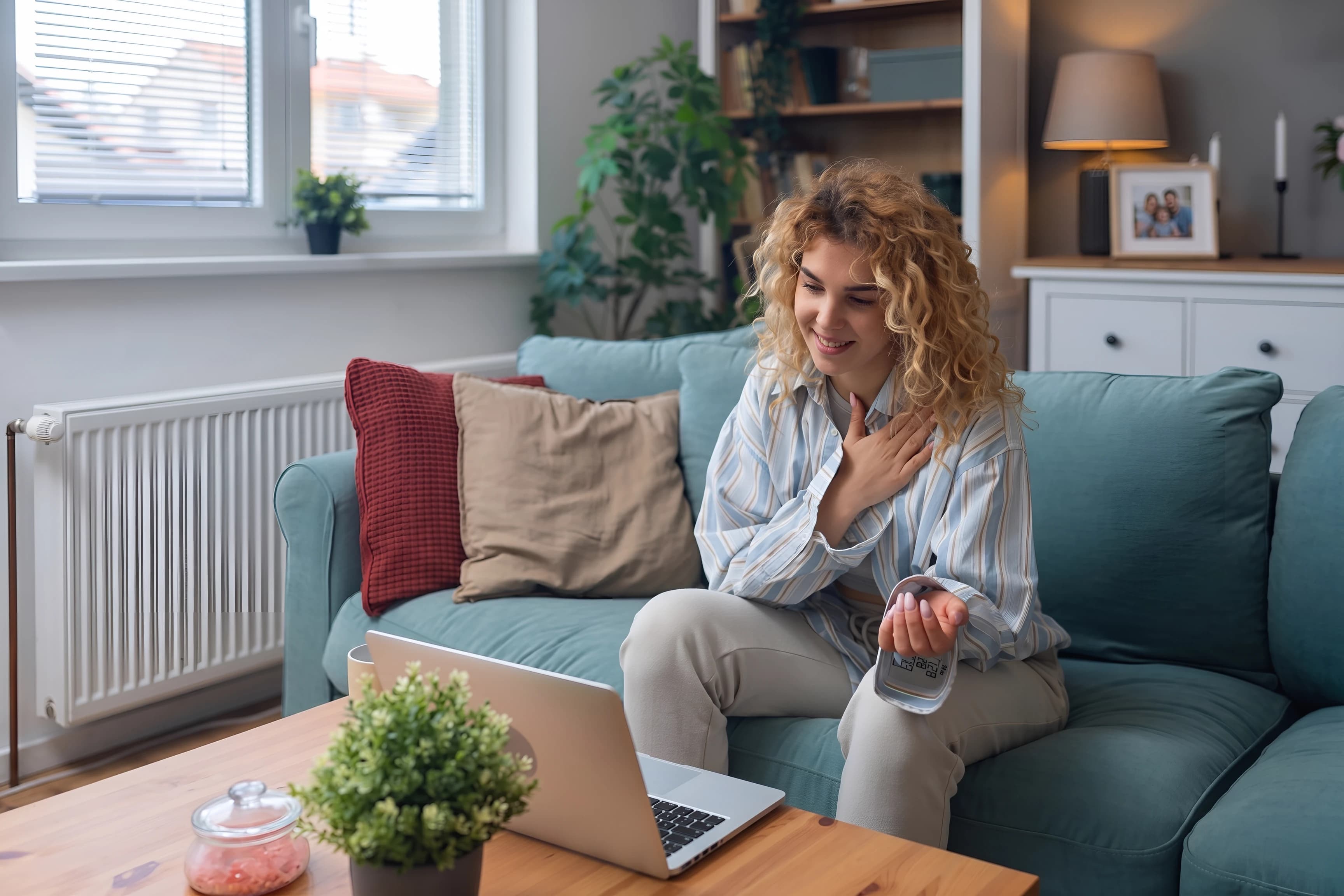 Smiling woman on a sofa checking her blood pressure beside a laptop